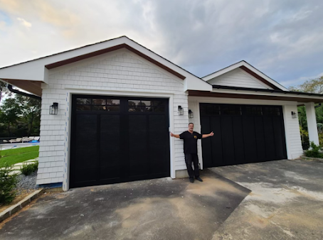 A man is standing in front of a white house with black garage doors.