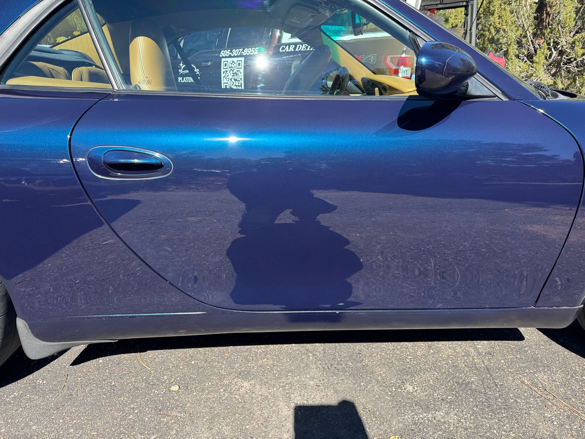 Black car's rear with shiny surface, under a canopy, car's tail light visible, sunny outdoor setting.