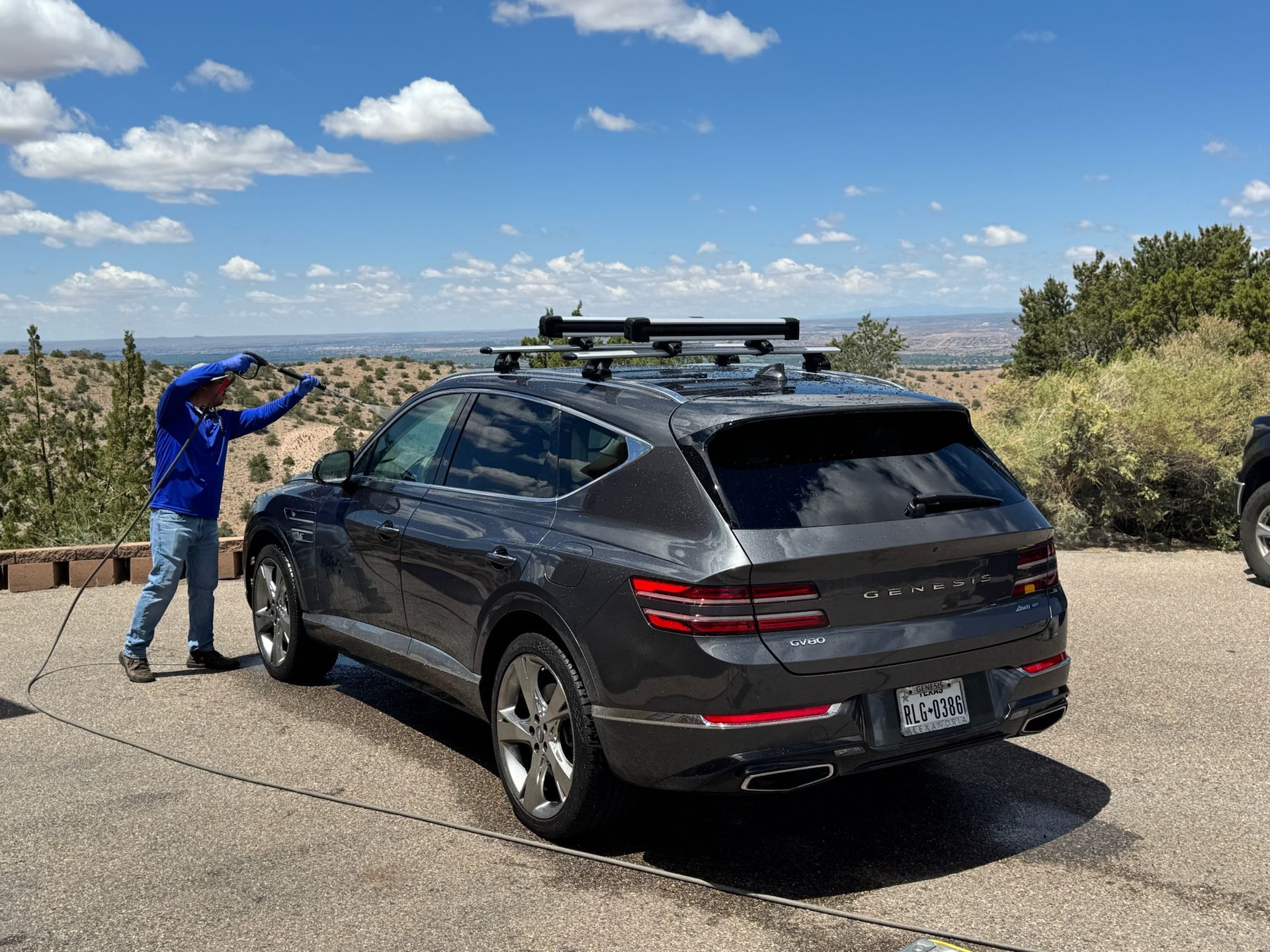 Person washing a gray SUV with a roof rack outdoors on a sunny day, with a blue sky and distant cityscape.