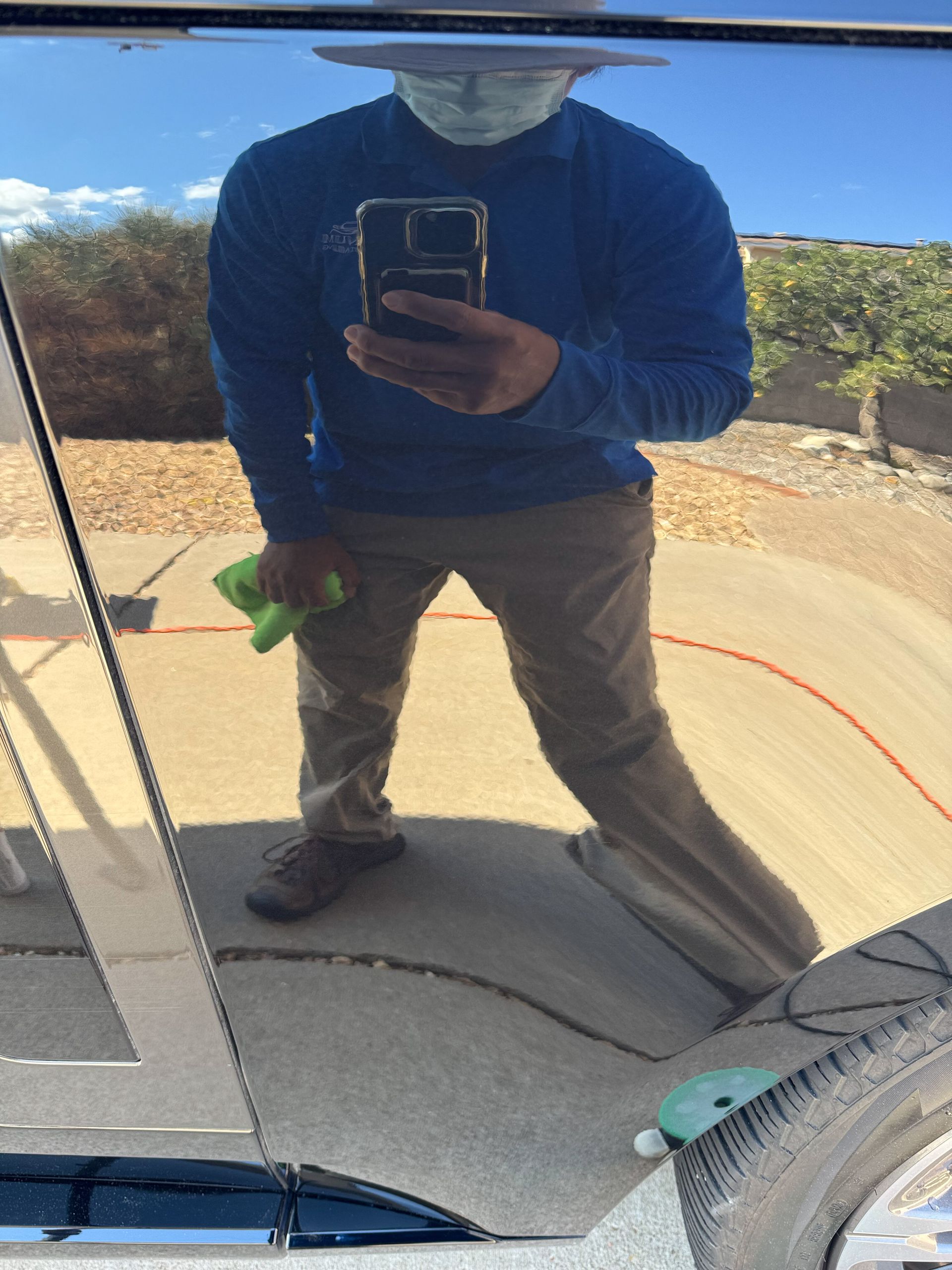 Person in blue shirt, tan pants, and mask holding a phone, reflected in car door, outdoors.