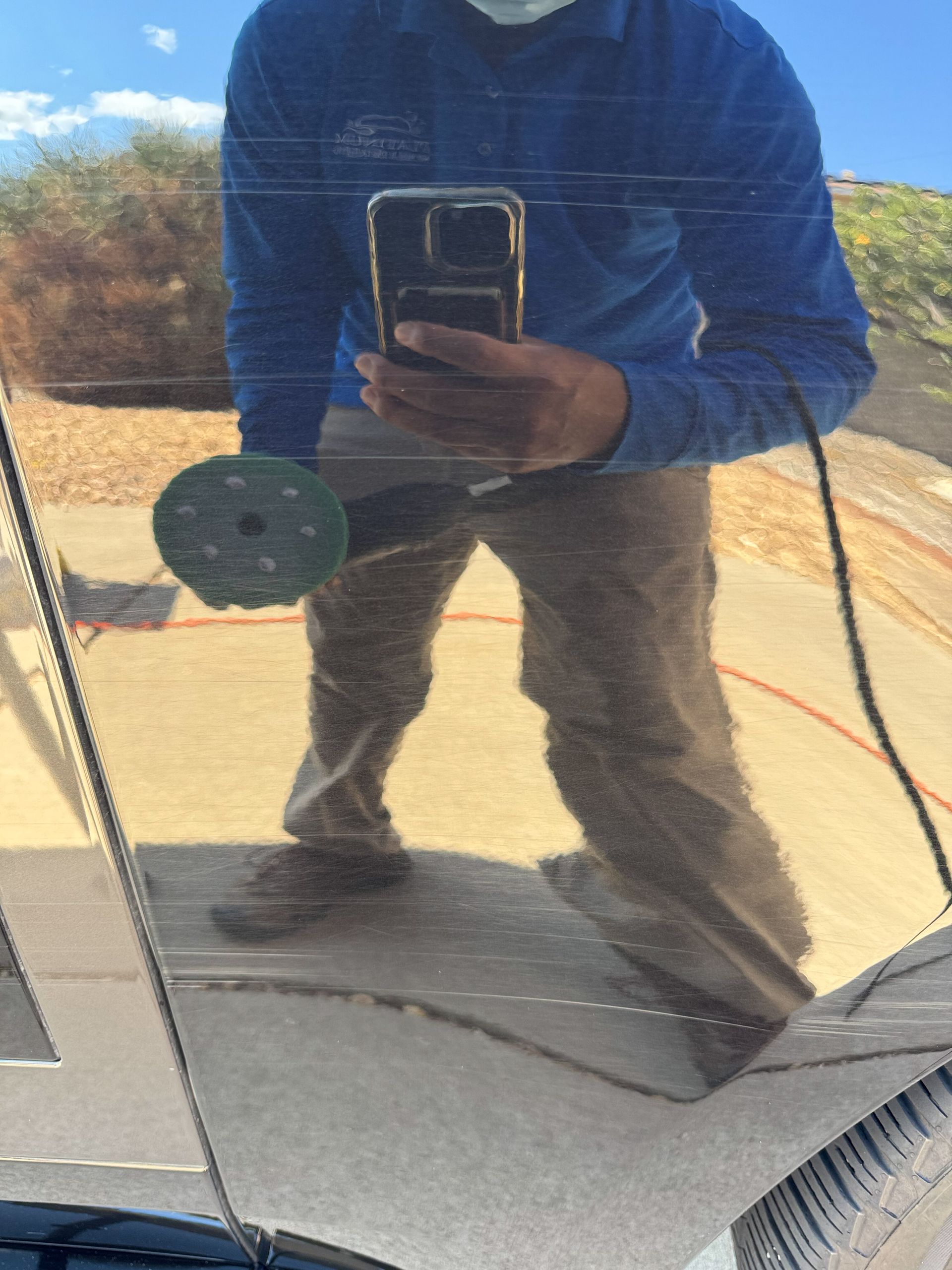Person polishing a car, reflected in the surface. Holding a buffer, wearing blue shirt and mask.
