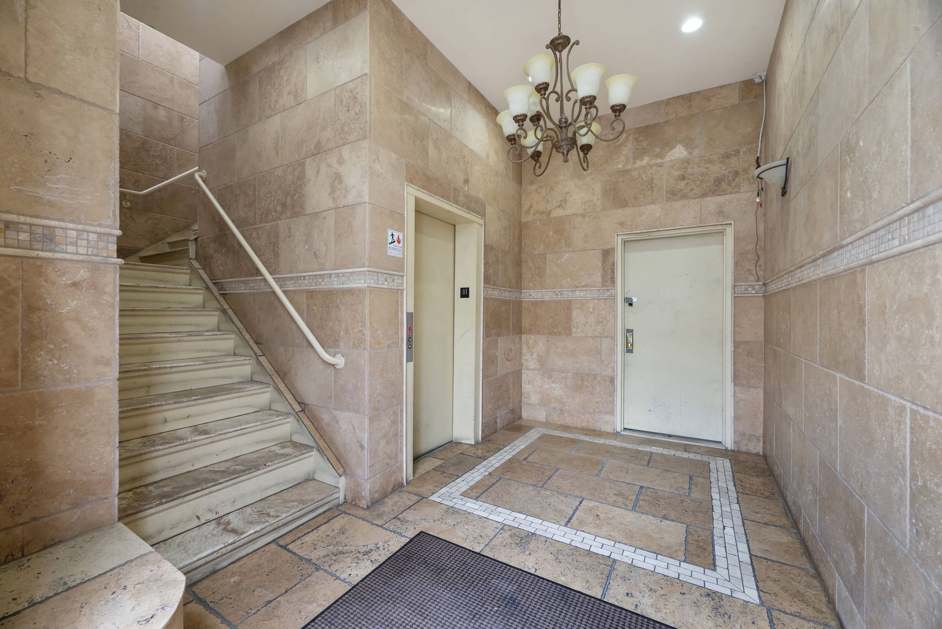A hallway with stairs leading up to an elevator and a chandelier hanging from the ceiling.