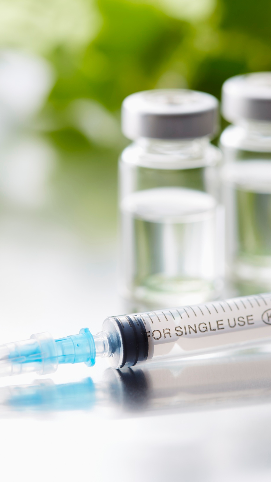 Syringe with needle next to two vials; the vials and syringe sit on a reflective surface.