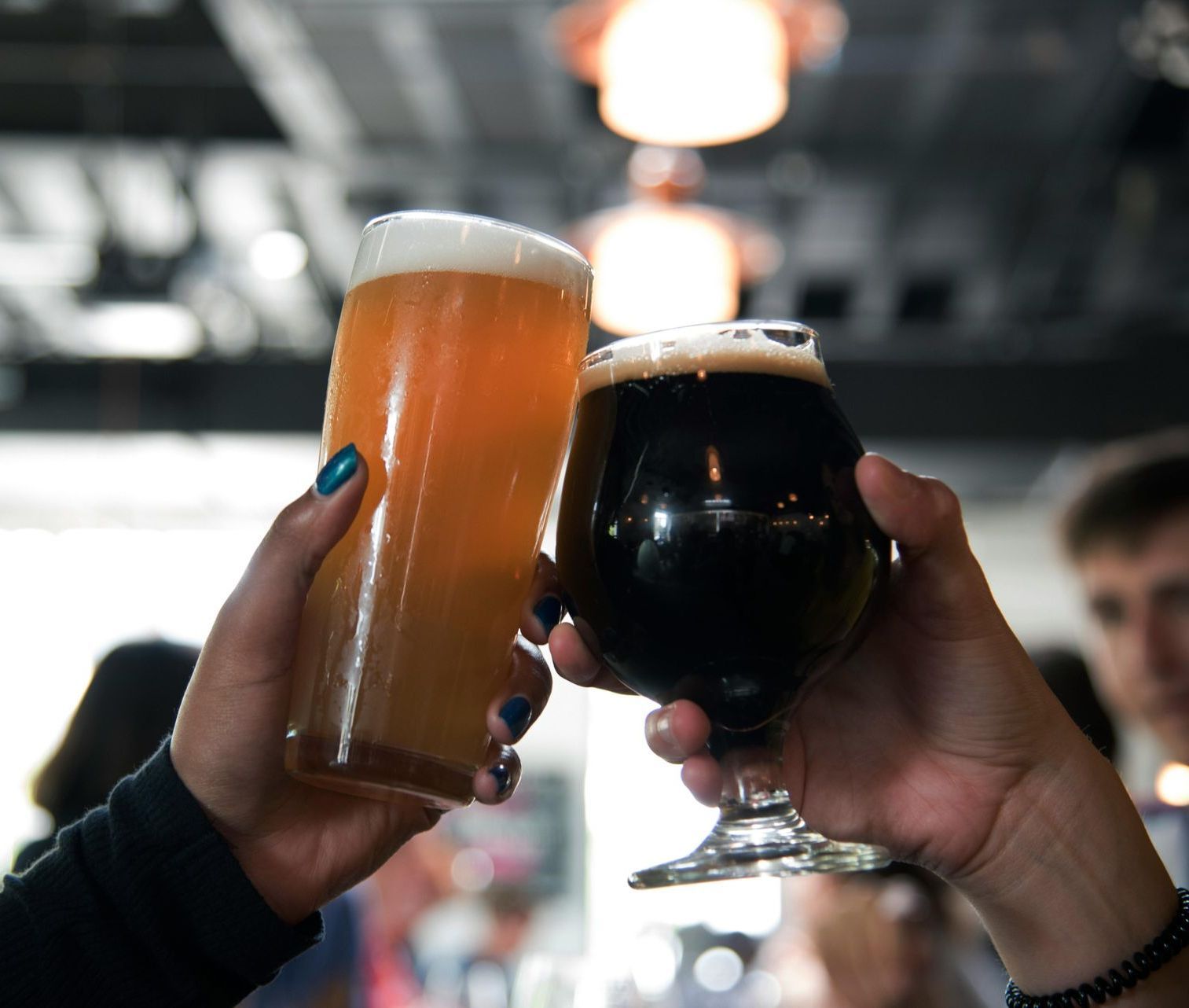 Two hands holding up beer glasses for a toast; one light and one dark, in a bar setting.