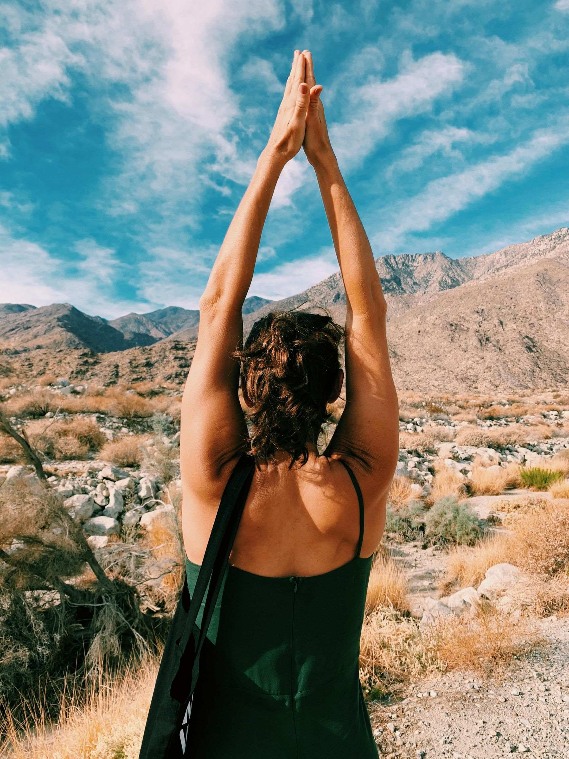 Woman in green dress, arms raised overhead, against a mountain landscape with blue sky.