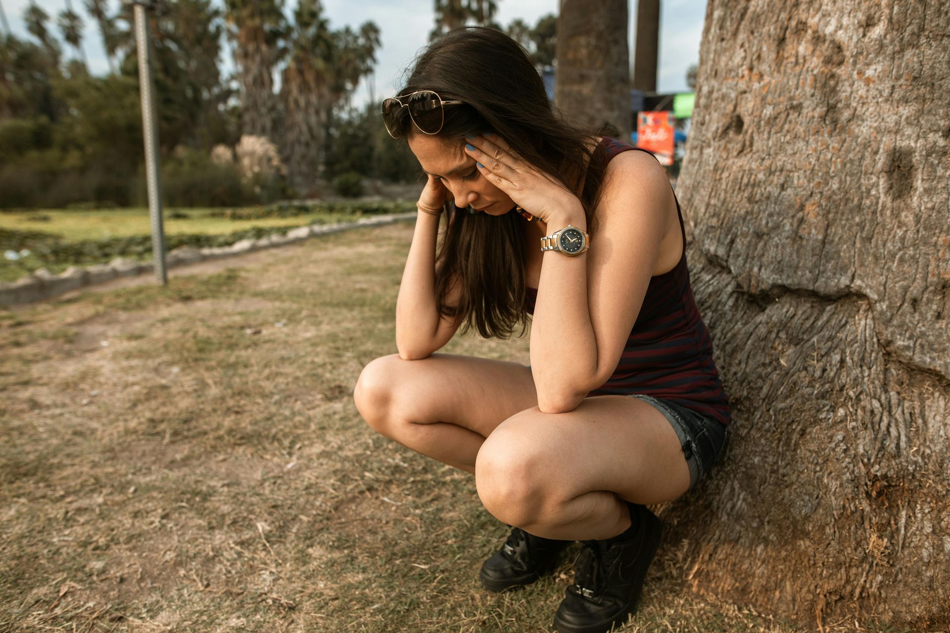 Woman squats in a park, touching her head with a pained expression. Leaning against a tree.