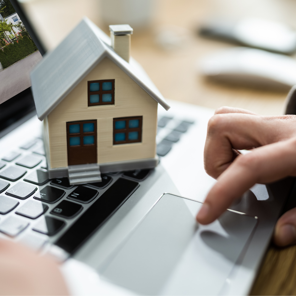 Person using laptop with a miniature house model on the keyboard, searching online for real estate.