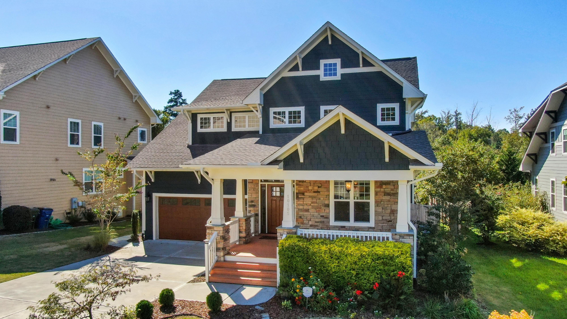 Two-story blue house with covered porch, brown garage door, and lush greenery.