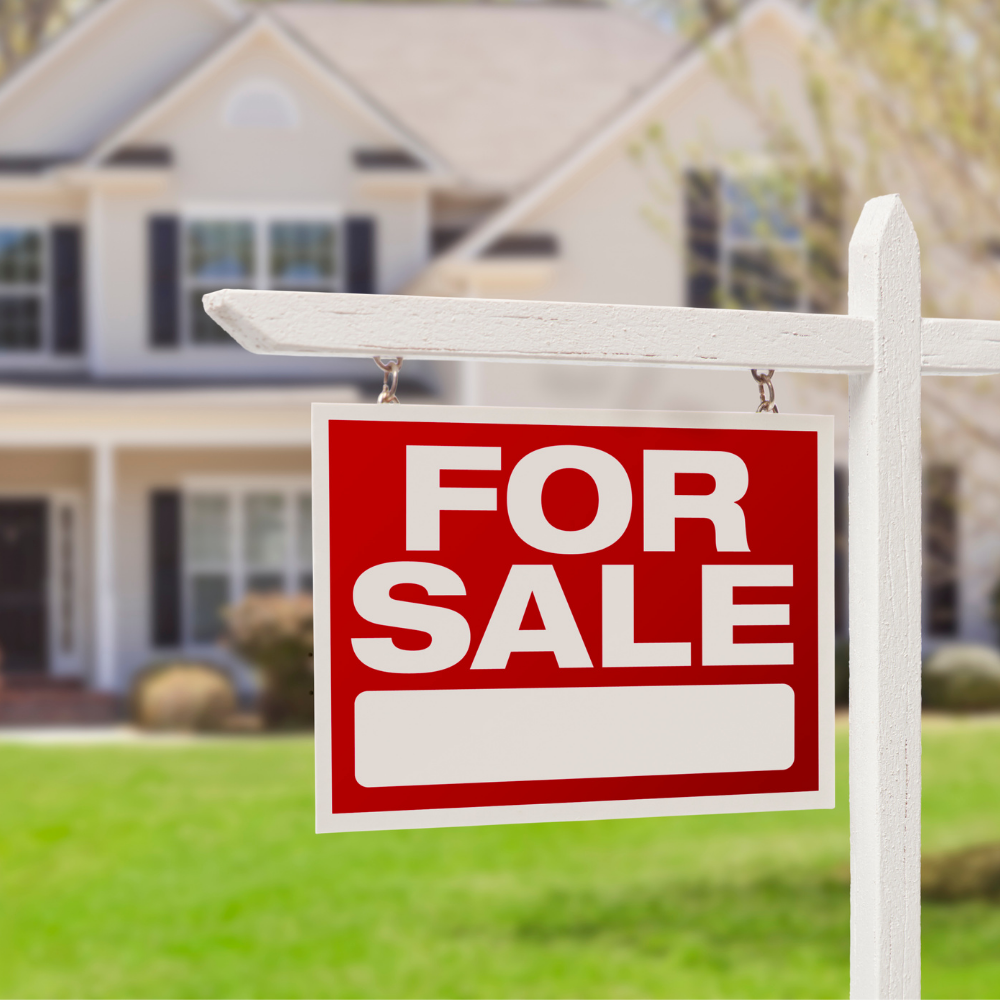 For sale sign in front of a light-colored house with a green lawn.