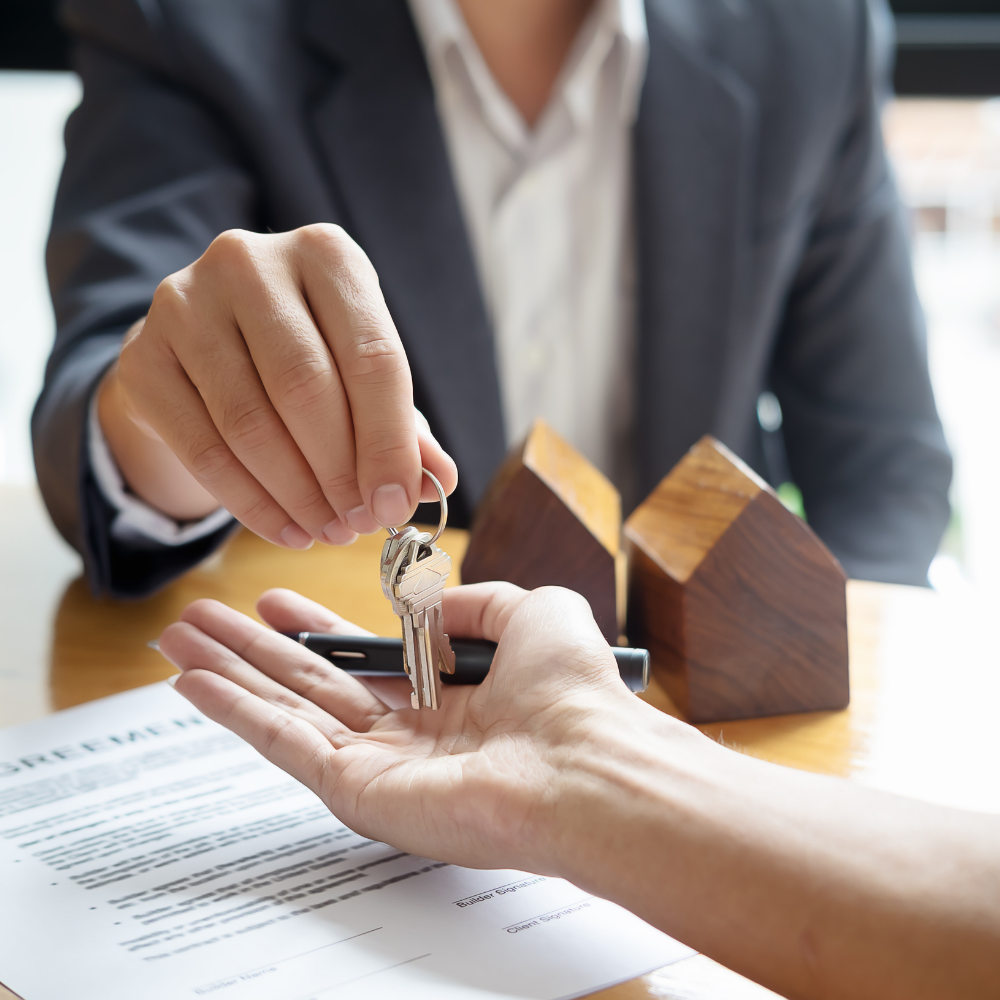 Person handing keys to another over a contract and model houses.