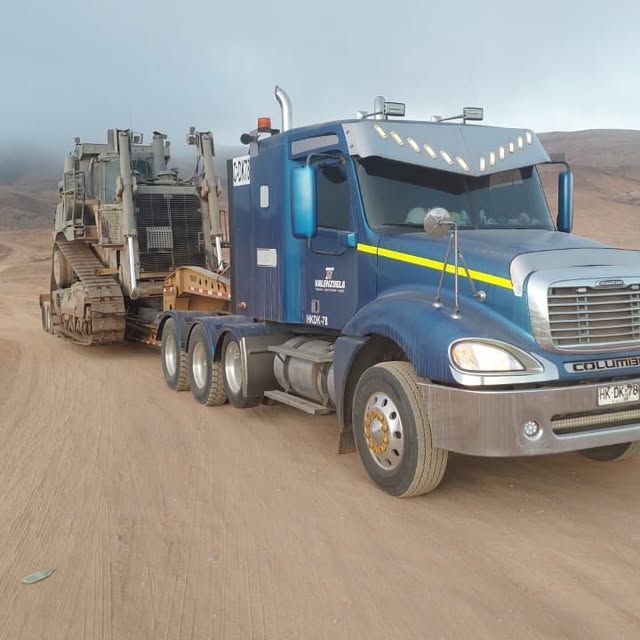 Un camión semirremolque azul transporta una excavadora por un camino de tierra.
