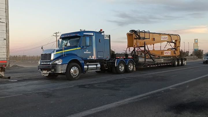 Un camión semirremolque azul circula por una autopista transportando una grúa.