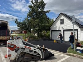 Paving crew laying asphalt driveway near a white garage on a sunny day. A Bobcat is visible.
