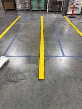 Yellow and blue floor markings in a warehouse, defining aisles and lanes on a concrete floor.