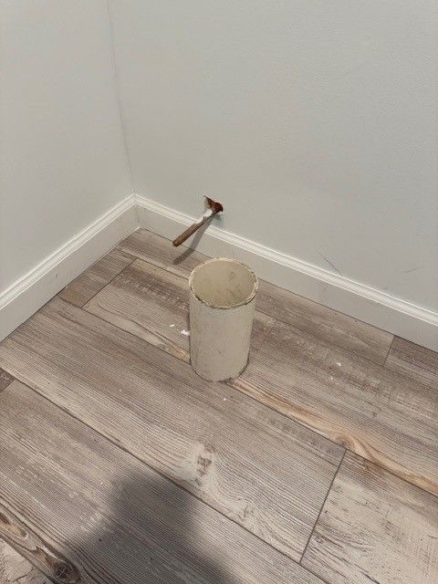 A white cylindrical pipe on wood-look flooring in a corner with white walls and molding; pliers on the wall
