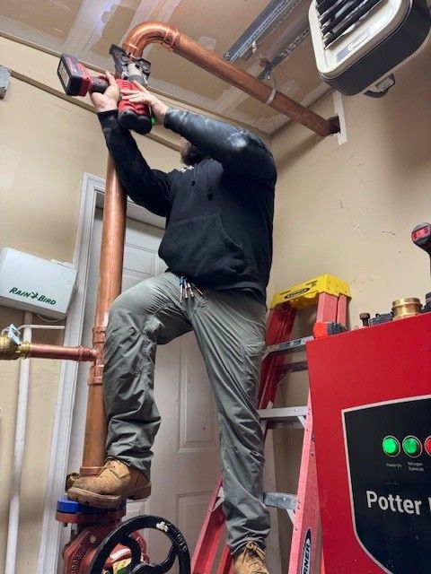 jared steen, owner of steen & son plumbing and heating standing on a ladder using a drill to assemble a commercial 3 inch line in mays landing, new jersey