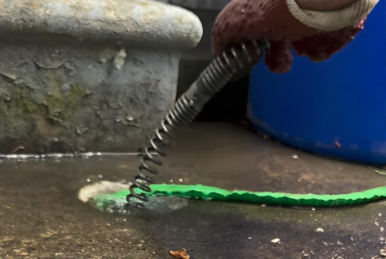 A technician from Steen & Son Plumbing and Heating wearing a glove uses a plumbing snake to unclog a drain, with water and debris visible on the surface in Mays Landing, New Jersey