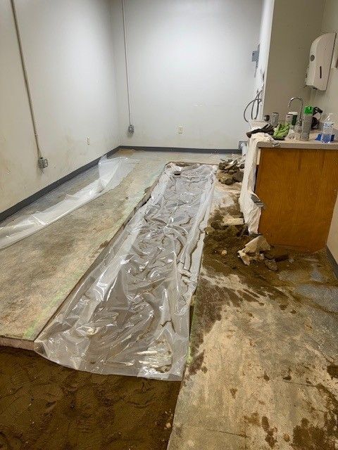 Room with plastic sheeting on a concrete floor, a waterlogged area, and a wooden cabinet being repaired by Steen & Son Plumbing and Heating in Mays Landing, NJ