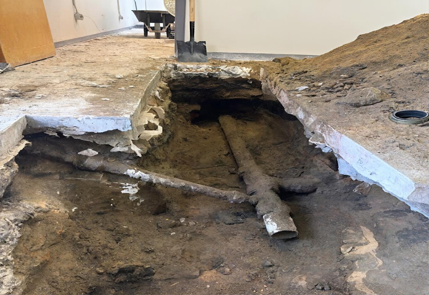Construction site floor with exposed plumbing pipes in a trench being fixed by Steen & Son Plumbing and Heating in Mays Landing, NJ. Brown dirt and gray concrete are visible, along with a partially visible wheelbarrow