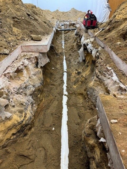 A long trench dug in dirt, with a white pipe running through the center. A worker operates a machine in the distance.
