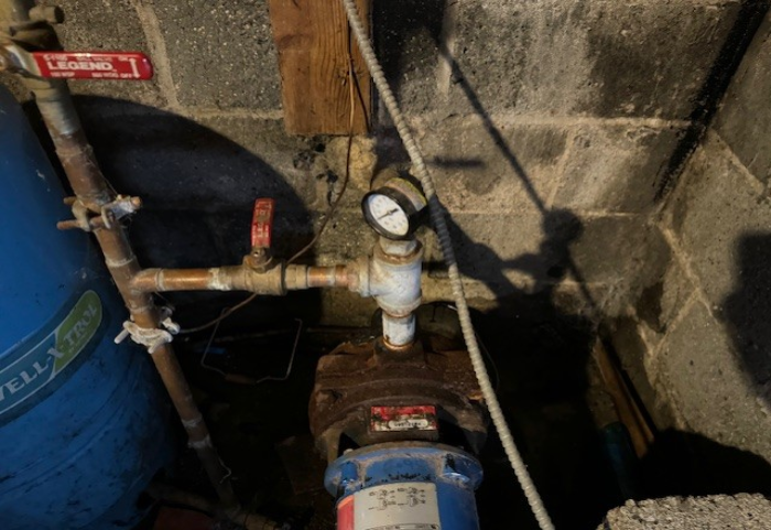 A well pump system with a blue tank, pipes, and a pressure gauge in a dark basement setting in Atlantic County, New Jersey being replaced by Steen & Son Plumbing and Heating