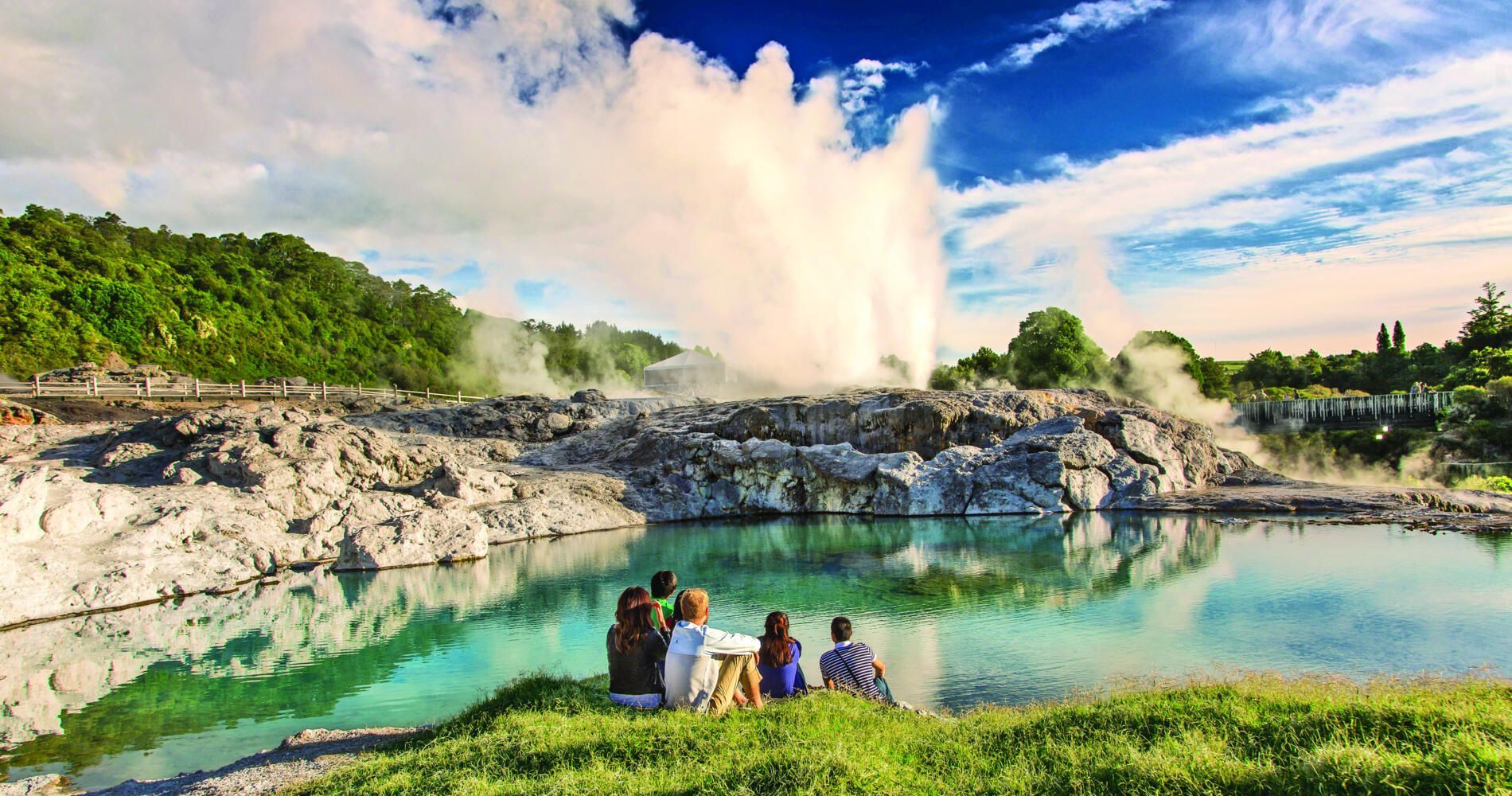 Um grupo de pessoas está sentado na grama perto de um lago observando a erupção de um gêiser.