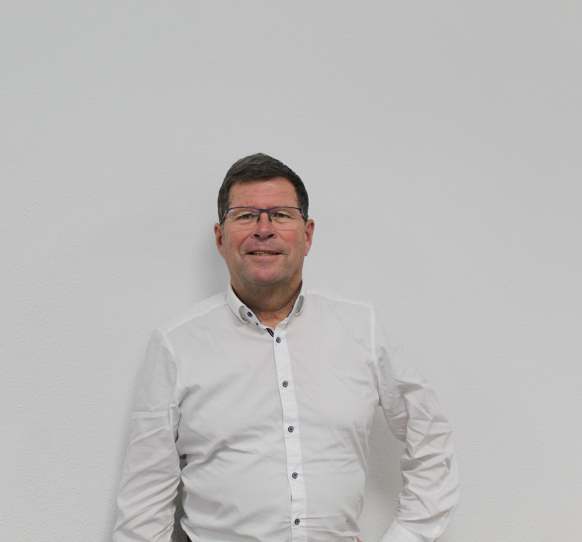 Een man met een bril en een wit overhemd staat voor een witte muur.