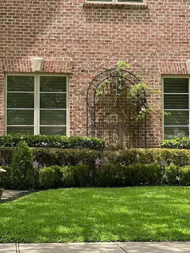 Red brick building with two windows and a rose arch; manicured hedges and lawn in the foreground.