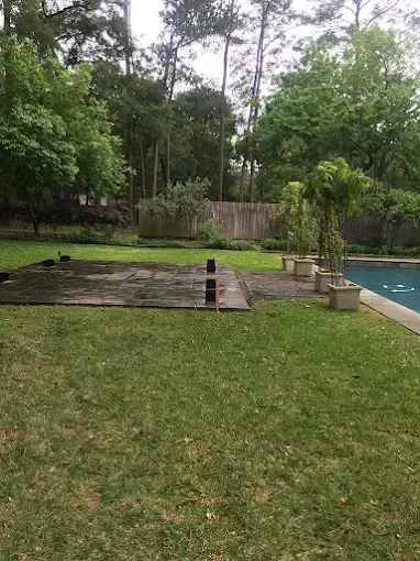 Green lawn and paved area next to a swimming pool, with trees and a wooden fence in the background.