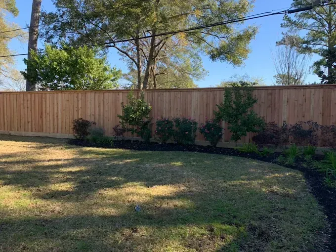 Wooden fence borders a backyard with green grass and planted bushes.