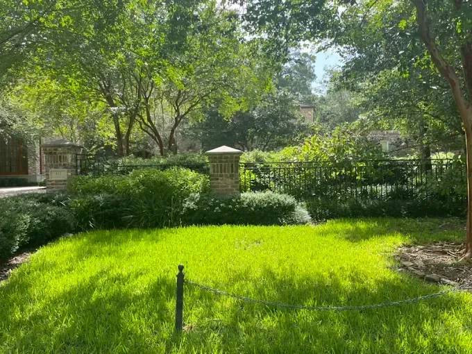 Green grassy yard with trees, bushes, and a brick post in a sunny outdoor setting.