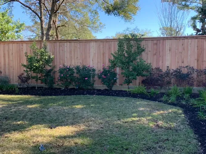 Wooden fence bordering a backyard with grass, flower beds, and trees.