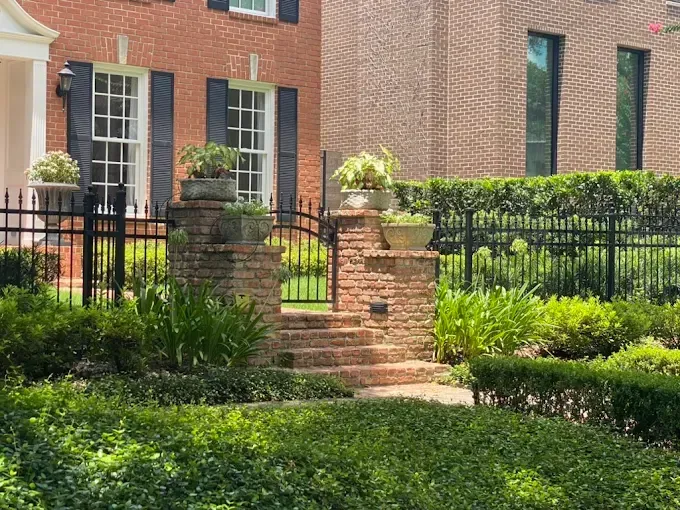 Brick and wrought-iron gate with steps, flanked by brick pillars with potted plants, in front of a red brick building.