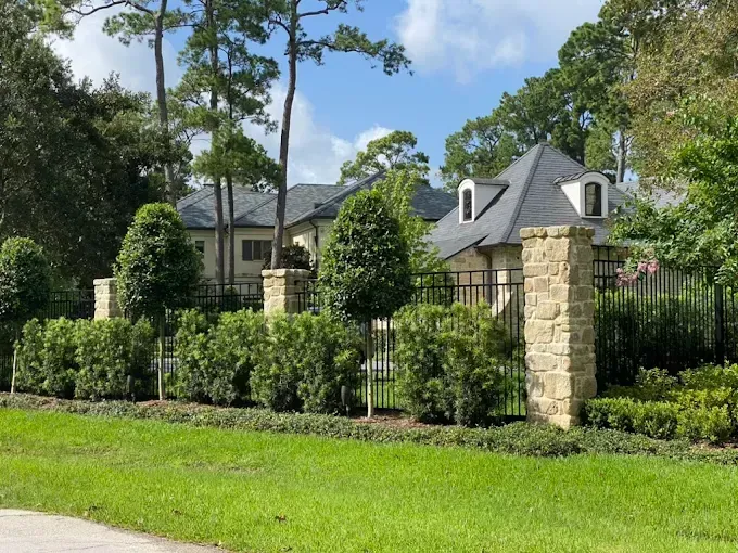 Stone-columned gate and fence enclose a large house with a gray roof, surrounded by trees and green lawn.