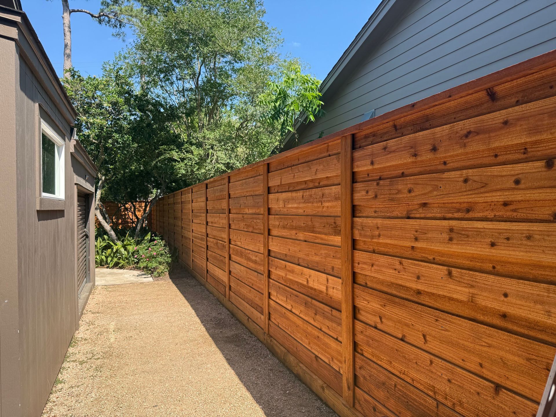 A wooden fence separates two buildings along a gravel path on a sunny day.