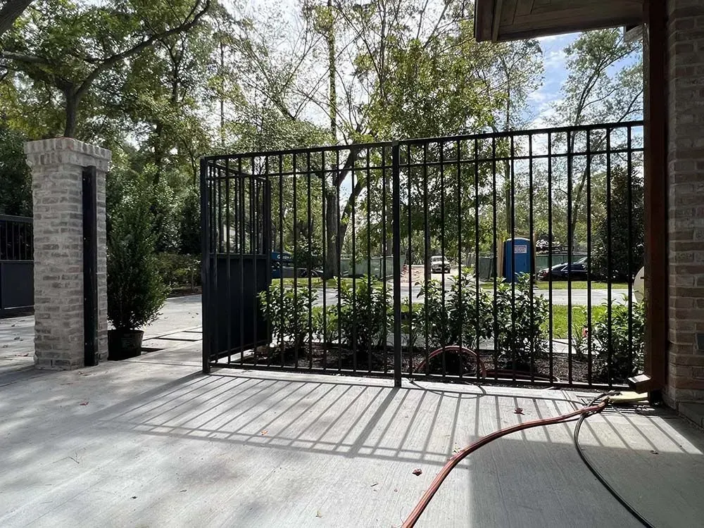 Black metal fence with a brick pillar, enclosing a garden with green bushes.