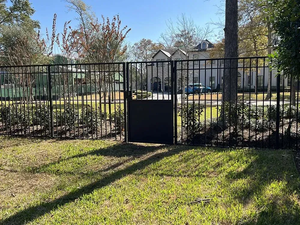 Black metal fence with a gate, in front of a yard with grass and bushes. A house can be seen in the background.
