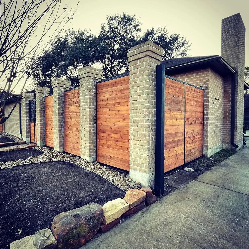Brick pillars frame a wooden fence in front of a brick house. Landscaping with rocks and dark soil is in view.