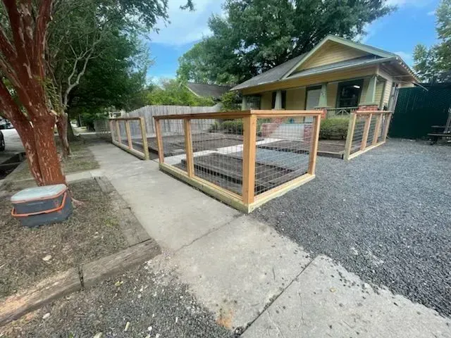 Raised garden beds with wire fencing sit on gravel next to a sidewalk and yellow house.