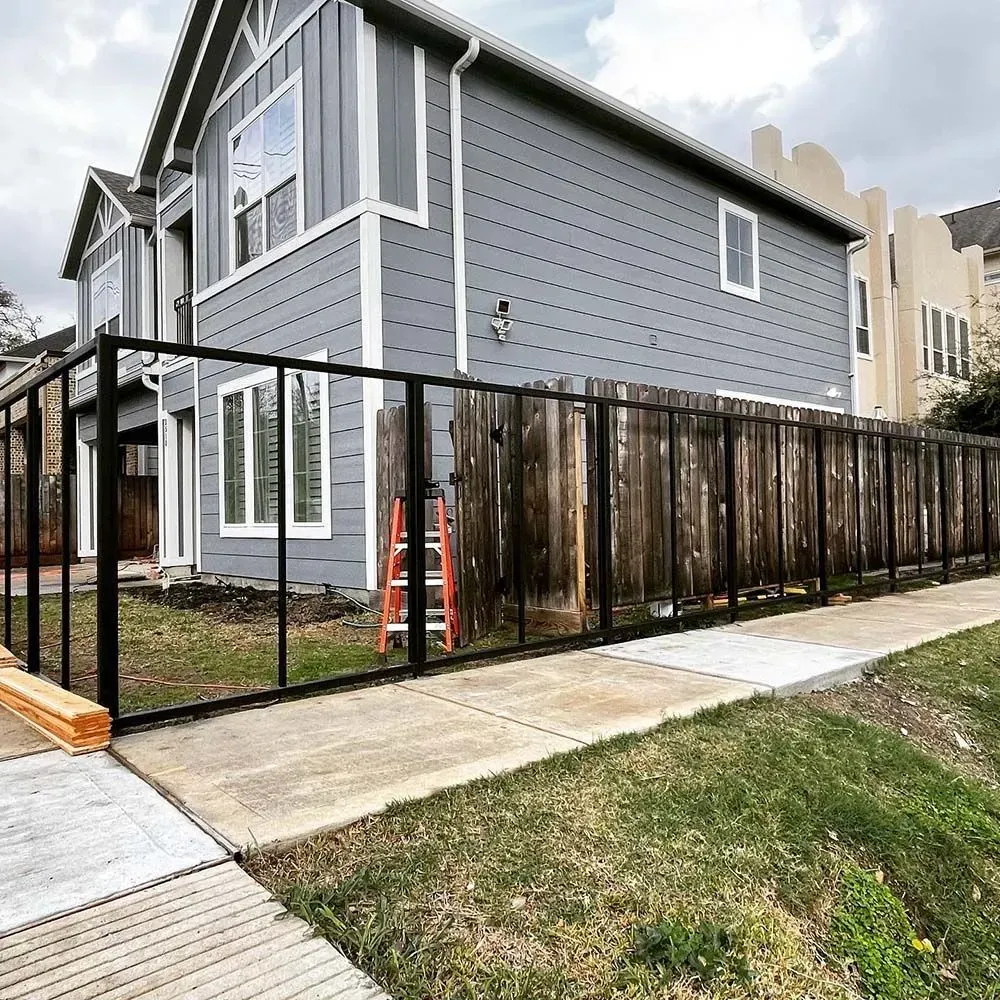 Black metal fence alongside a wooden fence next to a gray house, with a ladder leaned against the fence.