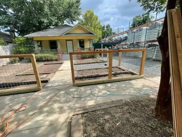 Wooden framed garden beds with wire fencing sit in front of a yellow house with a porch and walkway.