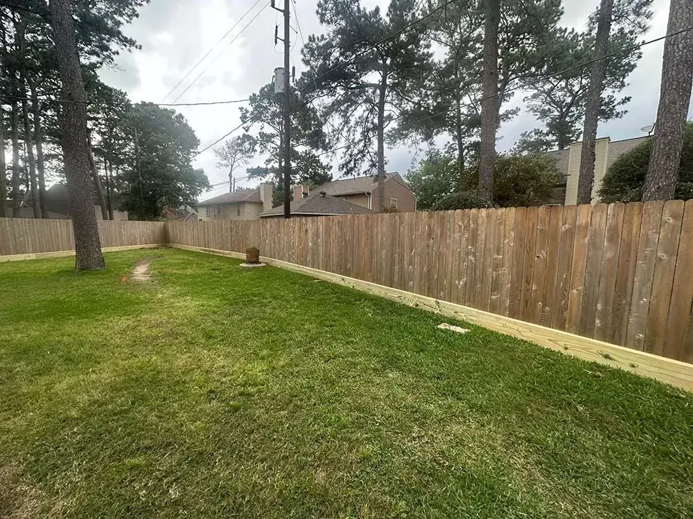 Backyard with wooden fence, green grass, and trees under a cloudy sky.