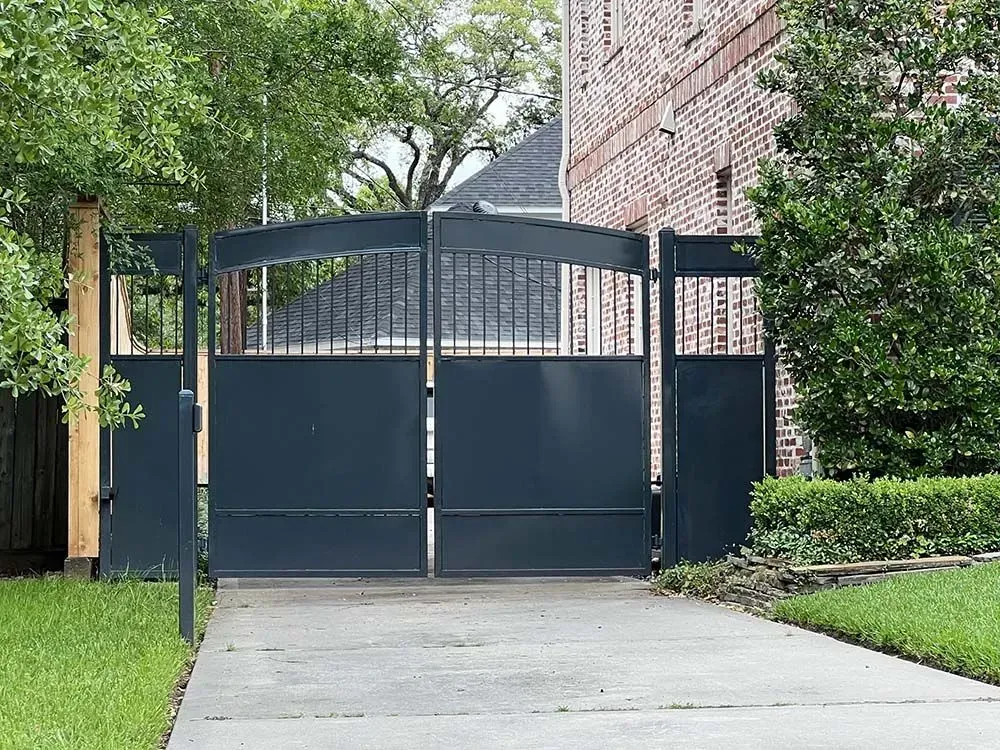 Dark blue metal driveway gate in front of a brick house with a concrete driveway and grass.