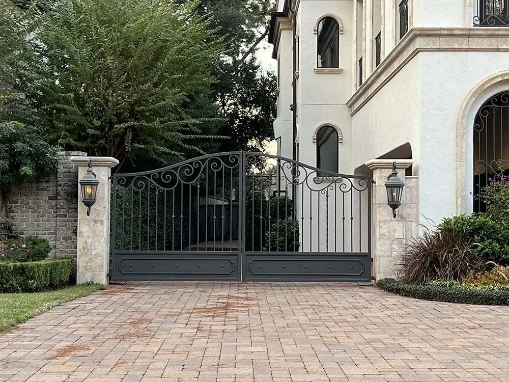 Iron gate at a home's entrance, brick driveway, stone columns with lanterns. White house with arched doorway.