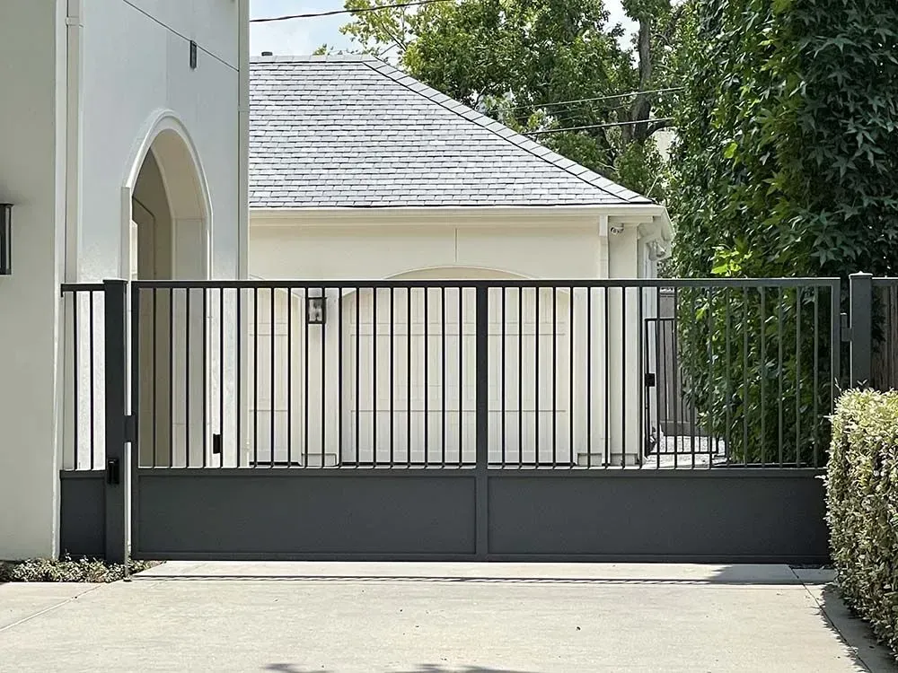 Gray metal driveway gate in front of a white house with a garage and green trees.
