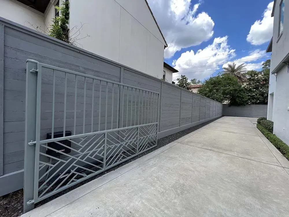 Gray gate, driveway, and fence against a backdrop of a white building and blue sky with clouds.