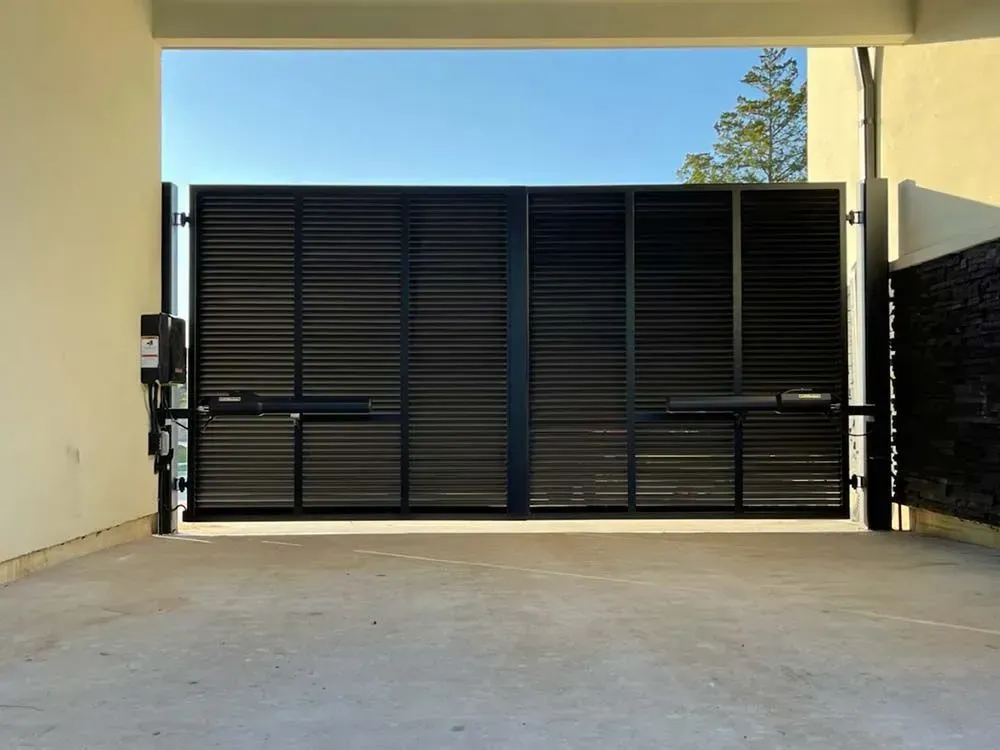 Black metal gate, closed, spans concrete driveway, between cream-colored walls, bright sky visible.