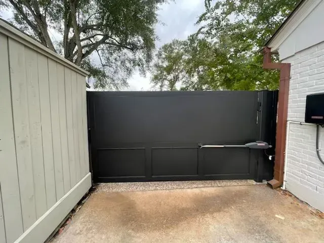 Black metal driveway gate, beige siding on the left, brick wall on the right, cloudy sky.