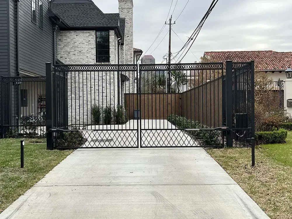 Black wrought-iron driveway gate. Concrete driveway in front of the gate. Houses in the background.