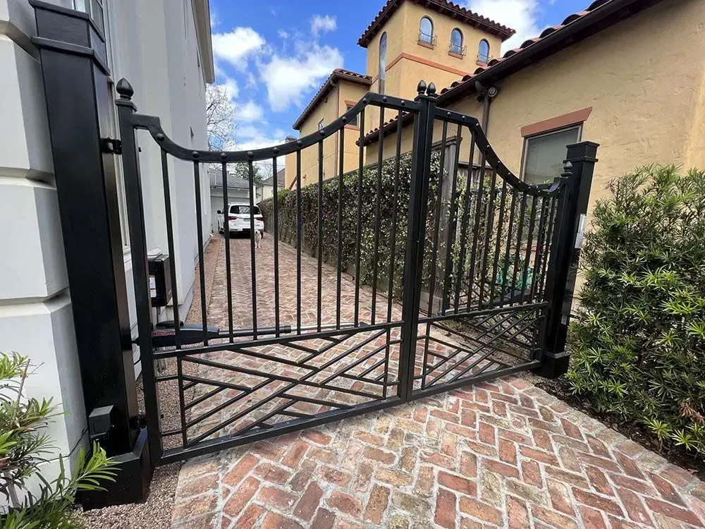 Black metal driveway gate with geometric design, brick paving, and a hedge on either side.
